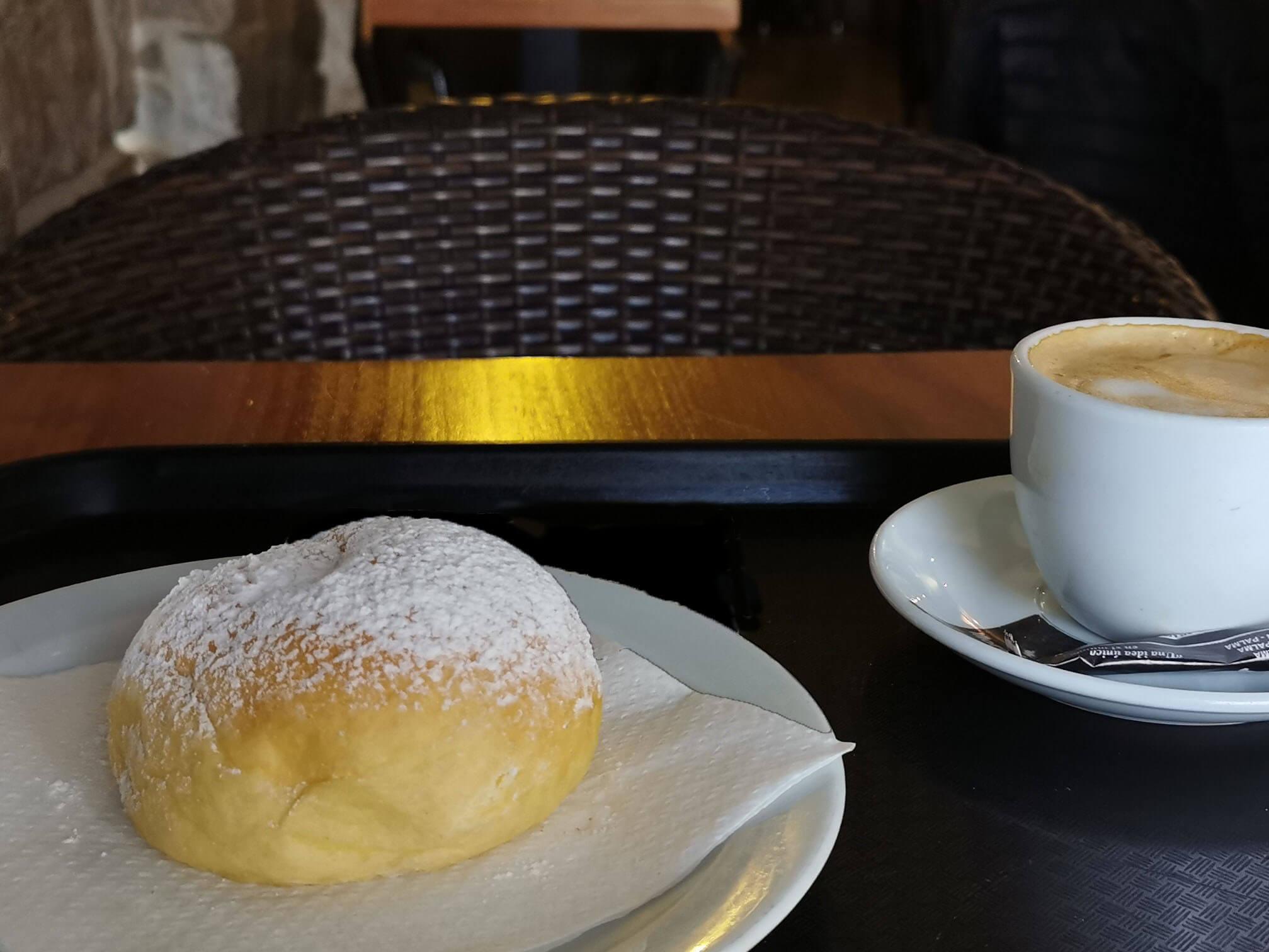 Ein typisches mallorquinisches Hefegebäck – Coca de Patata – auf einem Teller mit Puderzucker bestäubt, daneben eine Tasse Cappuccino in einem gemütlichen Café.