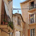Schmale Gasse in einer historischen Altstadt auf Mallorca mit traditionellen Balkonen, Pflanzen und einer alten Kirchenfassade im Hintergrund.