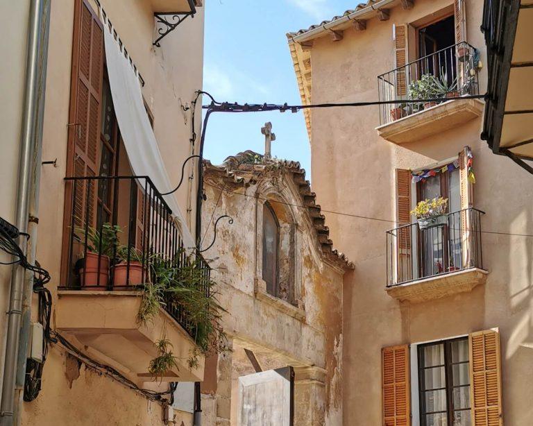 Schmale Gasse in einer historischen Altstadt auf Mallorca mit traditionellen Balkonen, Pflanzen und einer alten Kirchenfassade im Hintergrund.