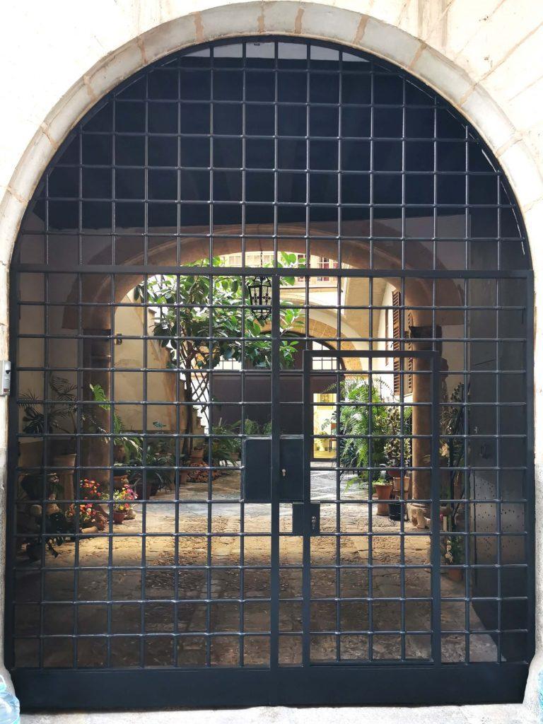 Blick durch ein schwarzes Gittertor in einen traditionellen mallorquinischen Innenhof mit Pflanzen, Steinboden und Rundbögen.