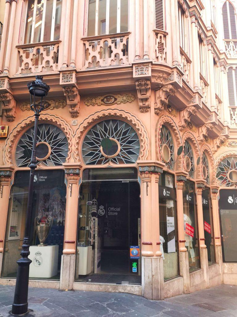 Historische Jugendstilfassade mit verschnörkelten Details und auffälligen Rundfenstern, in deren Erdgeschoss sich ein moderner Real-Madrid-Fanshop befindet.