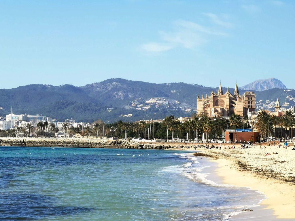 Blick auf die Kathedrale La Seu in Palma de Mallorca vom Strand aus, mit türkisblauem Meer, Palmen und dem Tramuntana-Gebirge im Hintergrund.