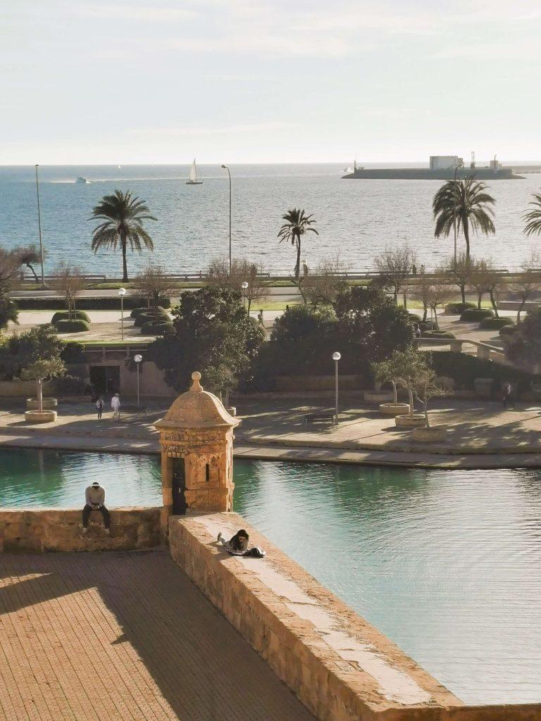 Blick auf das Meer, Palmen und einen alten Wachturm bei Palma de Mallorca mit entspannten Menschen in der Wintersonne.