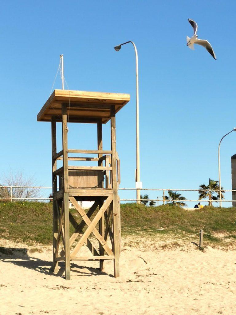 Hölzerner Rettungsturm am Sandstrand von Mallorca mit einer fliegenden Möwe im strahlend blauen Himmel.