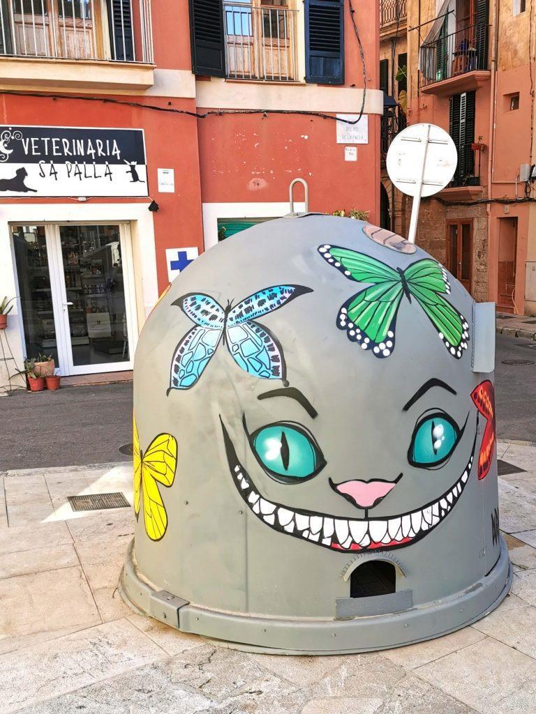 Bemalter Schutzcontainer für Straßenkatzen auf Mallorca, gestaltet mit einem bunten Katzen-Gesicht und Schmetterlingen – steht an einer Straßenecke vor einer Tierarztpraxis.