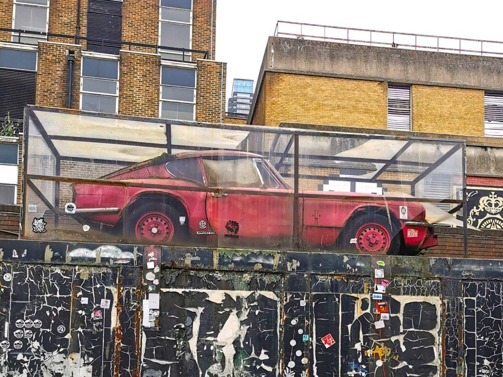 Altes rotes Auto hinter Plexiglas: Kunstinstallation auf einem Mauervorsprung in Shoreditch, London – Werk wird Banksy zugeschrieben.