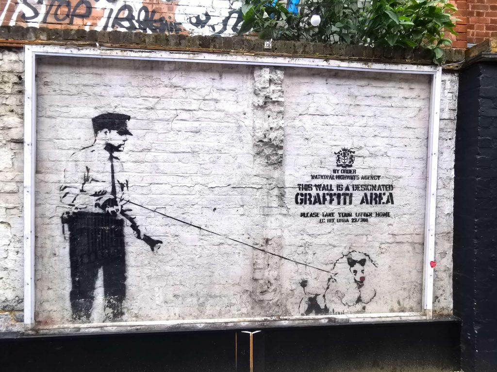 Banksy-Graffiti in London: Ein Polizist mit Hund vor einer Wand mit der Aufschrift ‚This wall is a designated graffiti area‘.