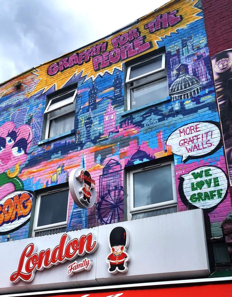 Buntes Streetart-Mural in Camden Town mit Londoner Sehenswürdigkeiten, Sprechblasen mit „Graffiti for the People“ und „We love Graff“, darunter ein Souvenirshop.