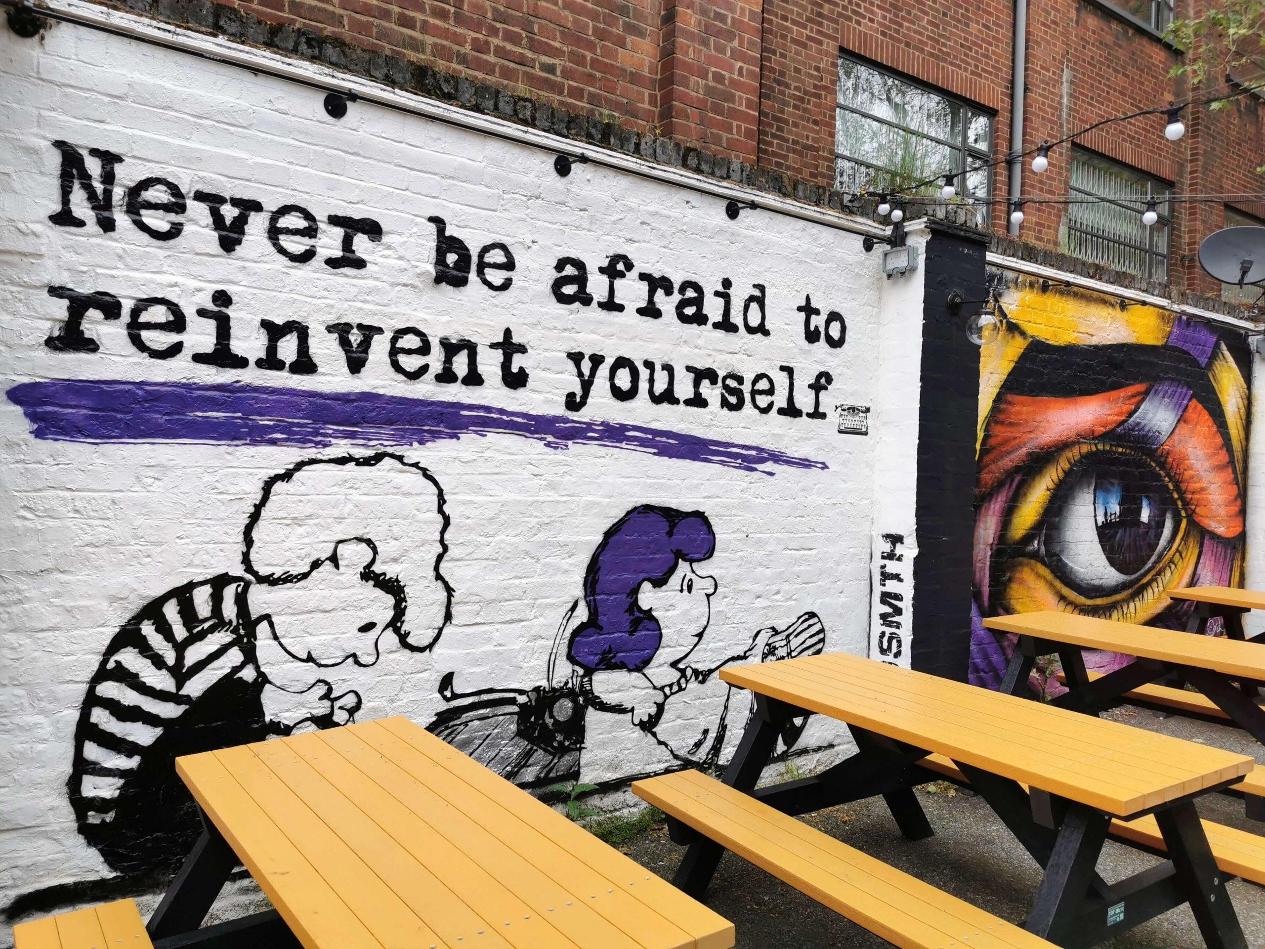 Streetart in London mit Peanuts-Zitat: ‚Never be afraid to reinvent yourself‘ – Comicfiguren, violette Details, Wandbild bei Picknickbänken