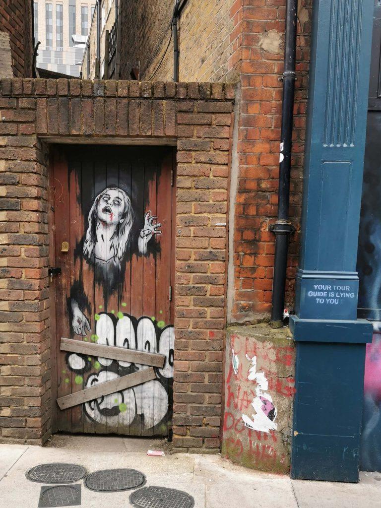 Streetart auf einer alten Holztür in London zeigt eine Zombie-Figur mit blutverschmiertem Mund. Daneben steht an einer Hauswand: ‚Your tour guide is lying to you‘.