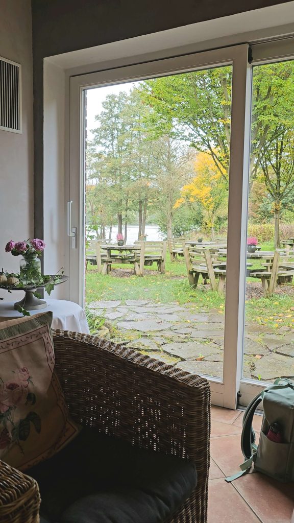 Blick aus dem Innenraum des Farchauer Mühlencafés auf die Gartenterrasse mit Holztischen und herbstlicher Aussicht auf den See.