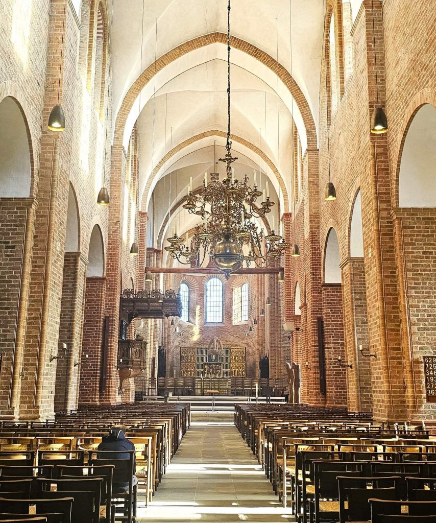 Blick in das Hauptschiff des Ratzeburger Doms mit vergoldetem Kronleuchter, hohen Backsteinbögen und warmem Sonnenlicht über den Kirchenbänken.