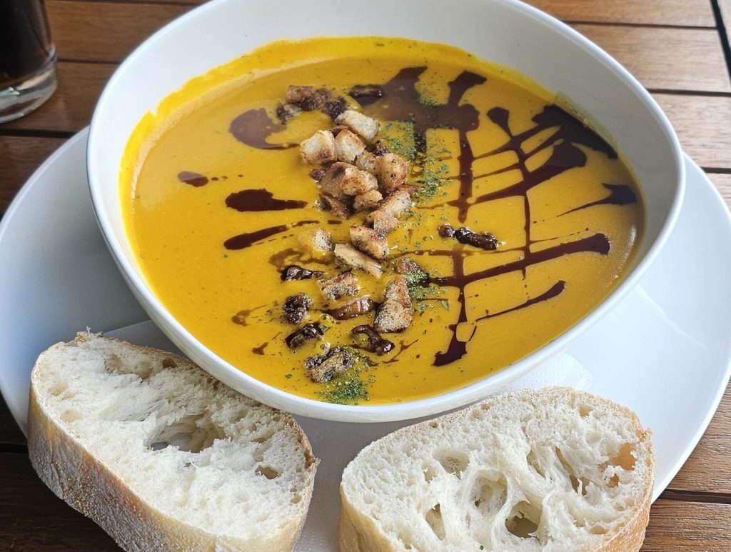 Nahaufnahme einer Kürbissuppe mit Croutons und Balsamico im Café in Ratzeburg, serviert mit frischem Baguette auf Holztisch.