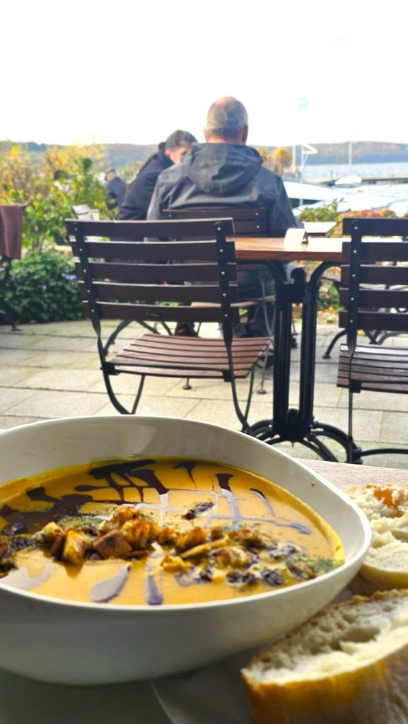 Schüssel Kürbissuppe mit Croutons und Balsamico, im Außenbereich eines Cafés am Ratzeburger See, mit Blick auf Wasser und Boote.