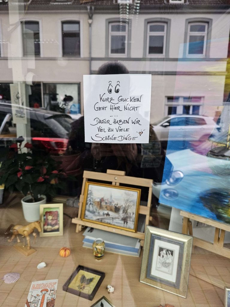 Schaufenster mit kleinen Kunstwerken, Bildern und Dekoartikeln in Ratzeburg. An der Scheibe hängt ein handgeschriebener Zettel mit der Aufschrift „Kurz gucken geht hier nicht – dafür haben wir viel zu viele schöne Dinge“.