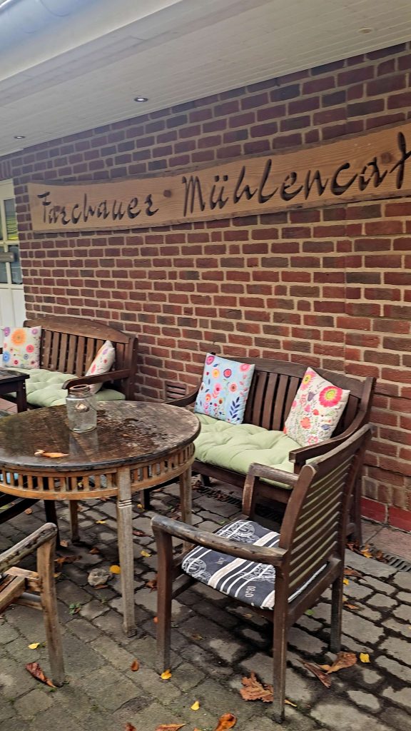 Gemütliche Terrasse des Fischhauer Mühlencafés mit Holztischen, Sitzbänken und bunten Blumenkissen an einer Backsteinwand.