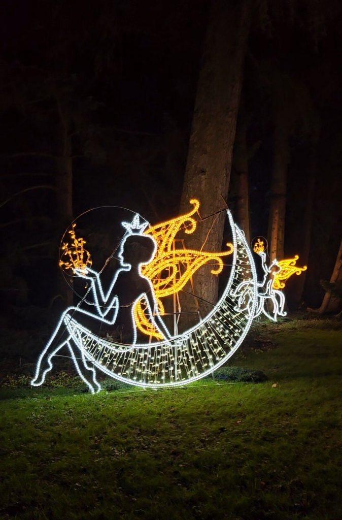 Lichtinstallation einer sitzenden Fee auf einer Mondsichel, beleuchtet in Weiß und Gold, am Waldrand der Insel Mainau.