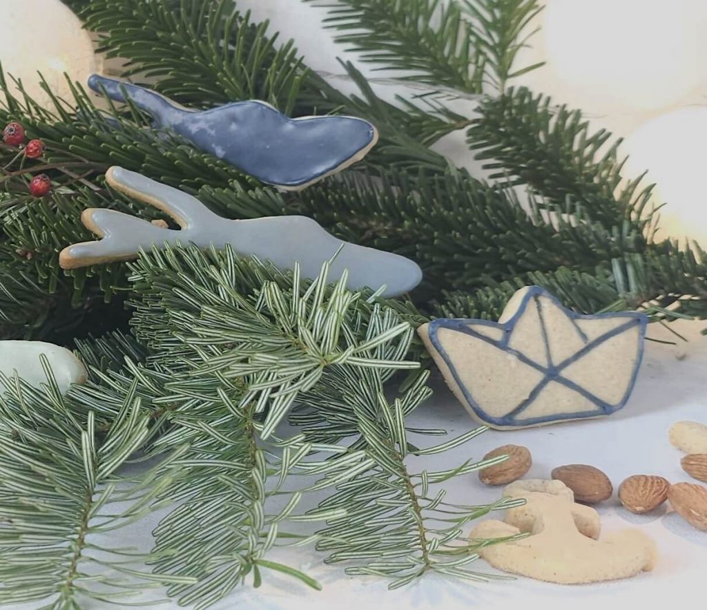 Weihnachtliche Kekse und Ausstecher in Form des Bodensees, Papierboot und eines Ankers zwischen Tannenzweigen und Mandeln.