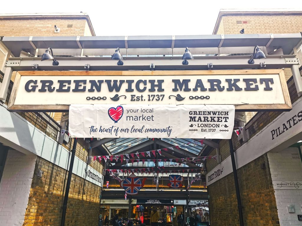 Eingangsschild des Greenwich Market in London mit Schriftzug und Markthalle im Hintergrund