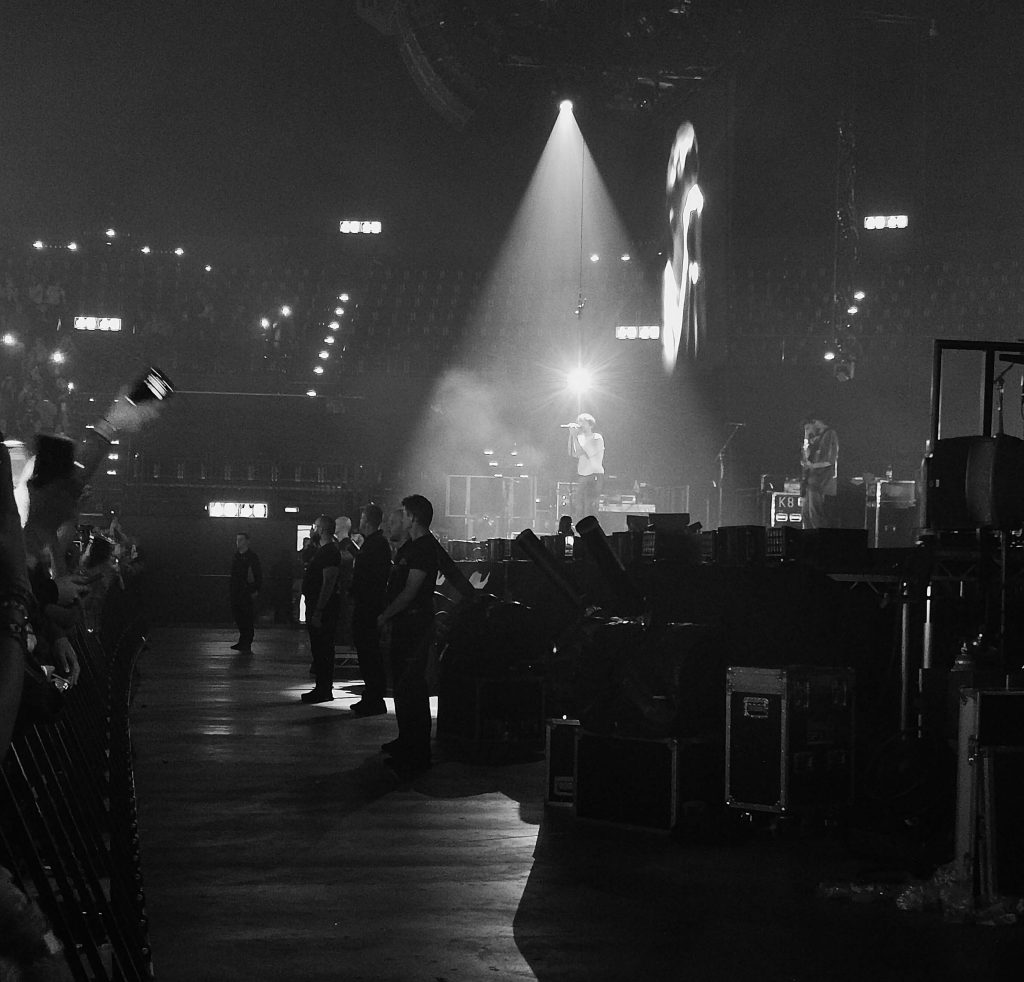 Schwarz-Weiß-Aufnahme eines Sängers unter Spotlicht auf einer Konzertbühne mit Publikum im Vordergrund