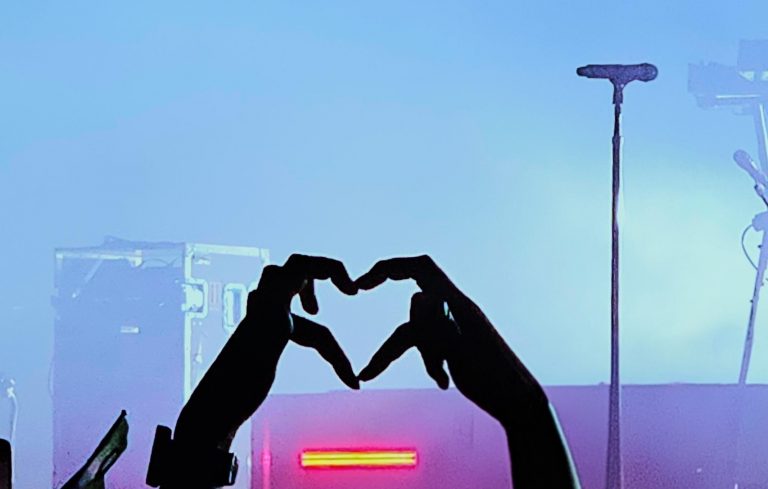 Silhouettenhände formen ein Herz vor einer beleuchteten Konzertbühne – Symbol für Gemeinschaft beim alleine auf ein Konzert gehen