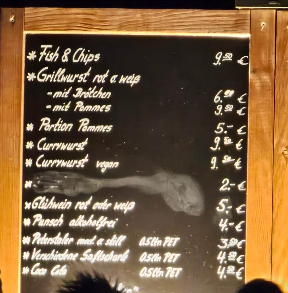 Preistafel mit Fish & Chips, Grillwurst, Pommes und Currywurst beim Christmas Garden Mainau in der Saison 2025/2026